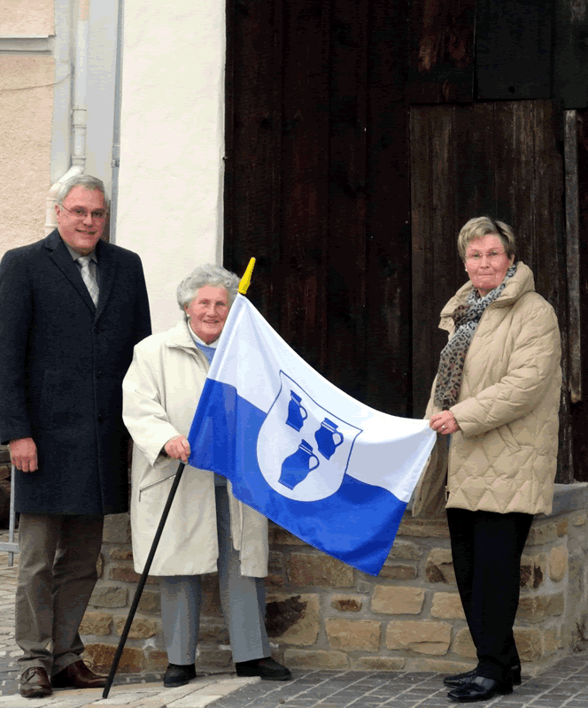 Der Stadtbürgermeister Michael Thiesen überreicht Frau Marga Gerhards die Stadtfahne im Beisein Ihrer Schwester Liselotte Herzmann